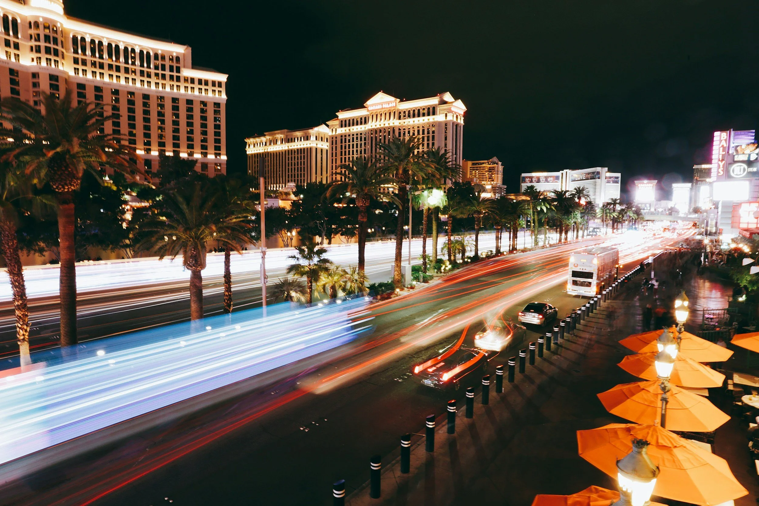 Nighttime view of the Las Vegas Strip with illuminated hotels and casinos, palm trees lining the street, and streaks of light from moving vehicles.
