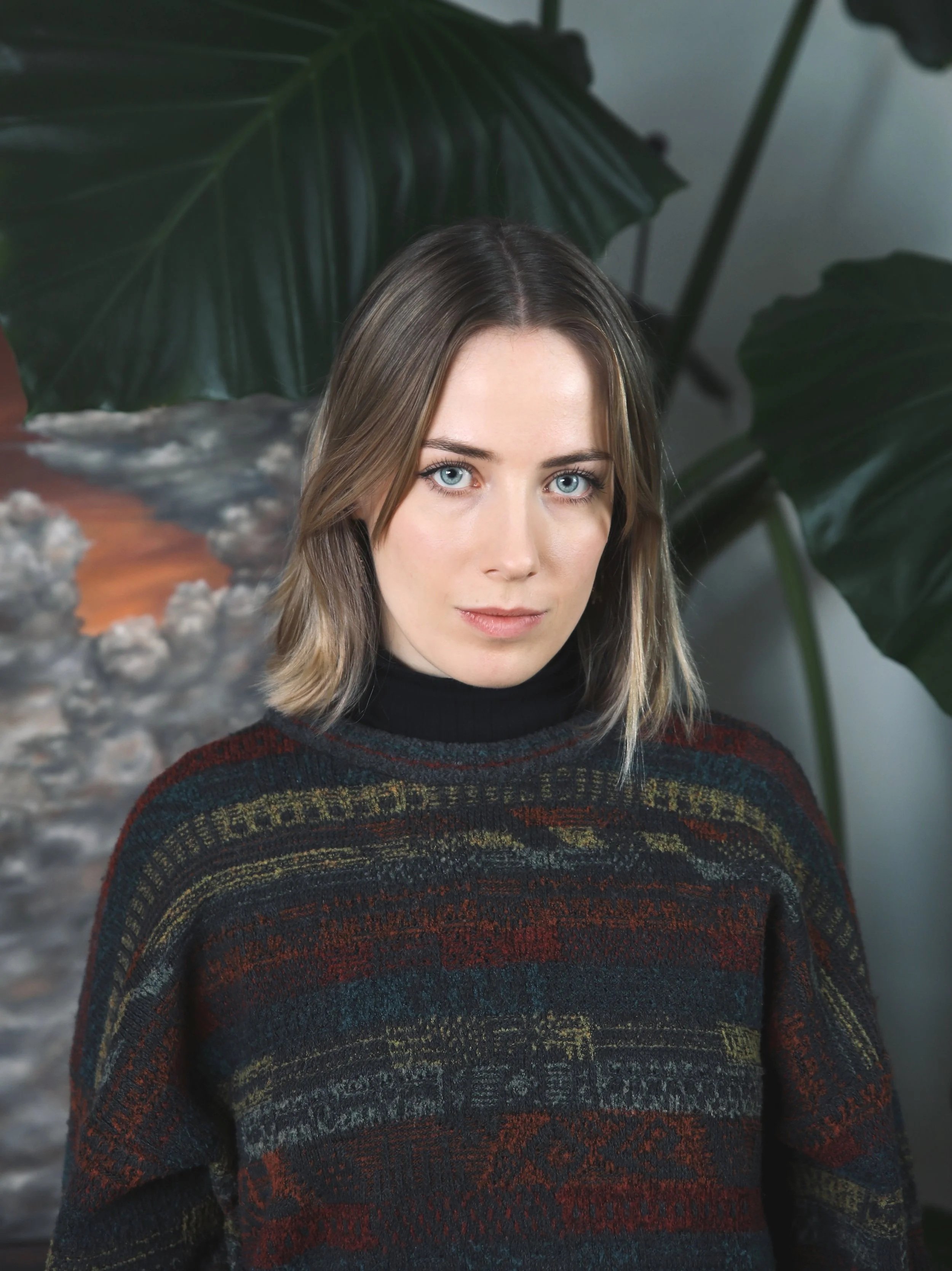 A woman with shoulder-length light brown hair, blue eyes, wearing a dark turtleneck and a multicolored patterned sweater, standing in front of large green leaves and a textured artwork.