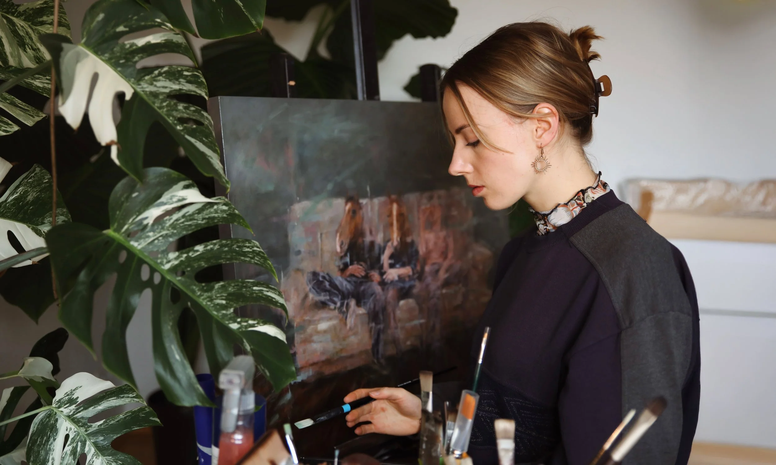A woman with brown hair in a bun, wearing a black top, patterned scarf, and decorative earrings, looks down while working at an art station surrounded by paintbrushes and painting supplies. There is a plant with large, green and white variegated leaves in the foreground, and a painting of a person on a brick wall in the background.