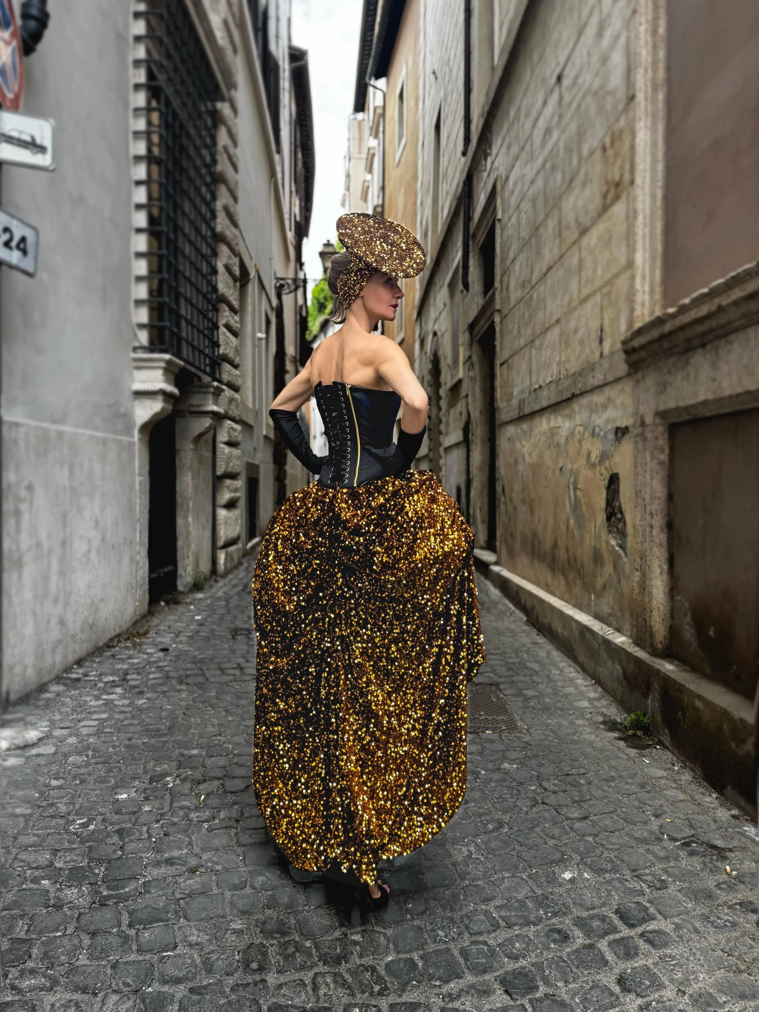 Donna di spalle in un vicolo con corsetto nero, gonna di paillettes dorate e cappello decorato