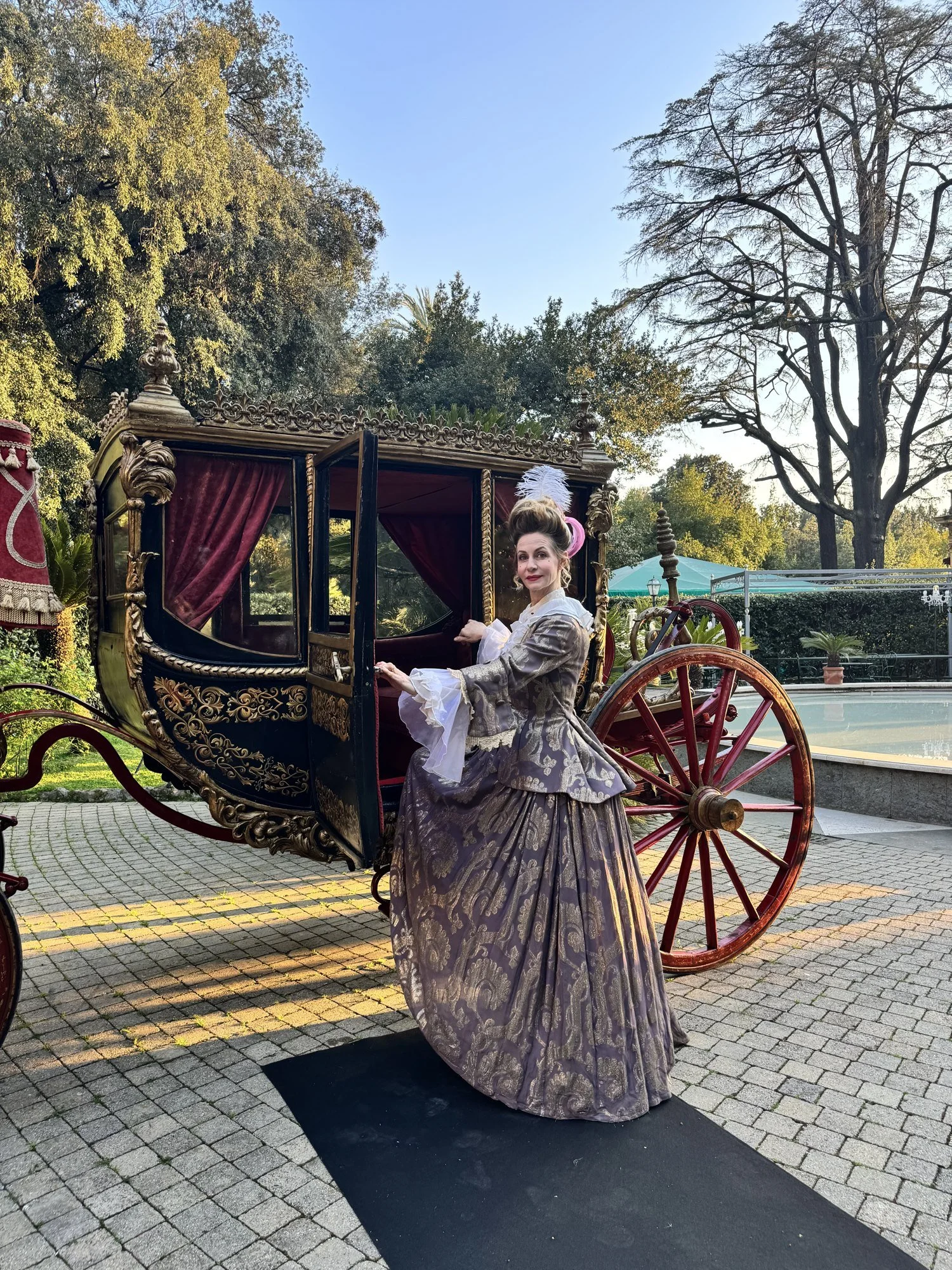 Donna in abito baroDonna in abito barocco accanto a una carrozza d'epoca tracco accanto a una carrozza d'epoca tra gli alberi, creazione Nice To Dress Youonna in abito barocco accanto a una carrozza d'epoca tra gli alberi, creazione Nice To Dress You