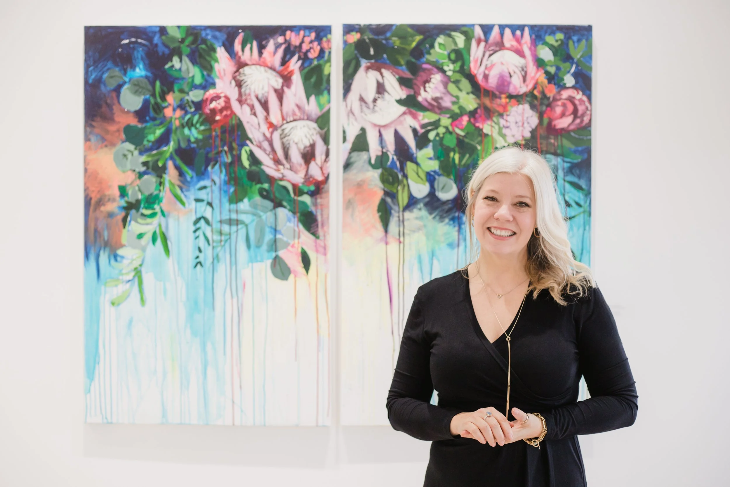 A woman smiling in front of a colourful abstract floral painting at an art gallery.