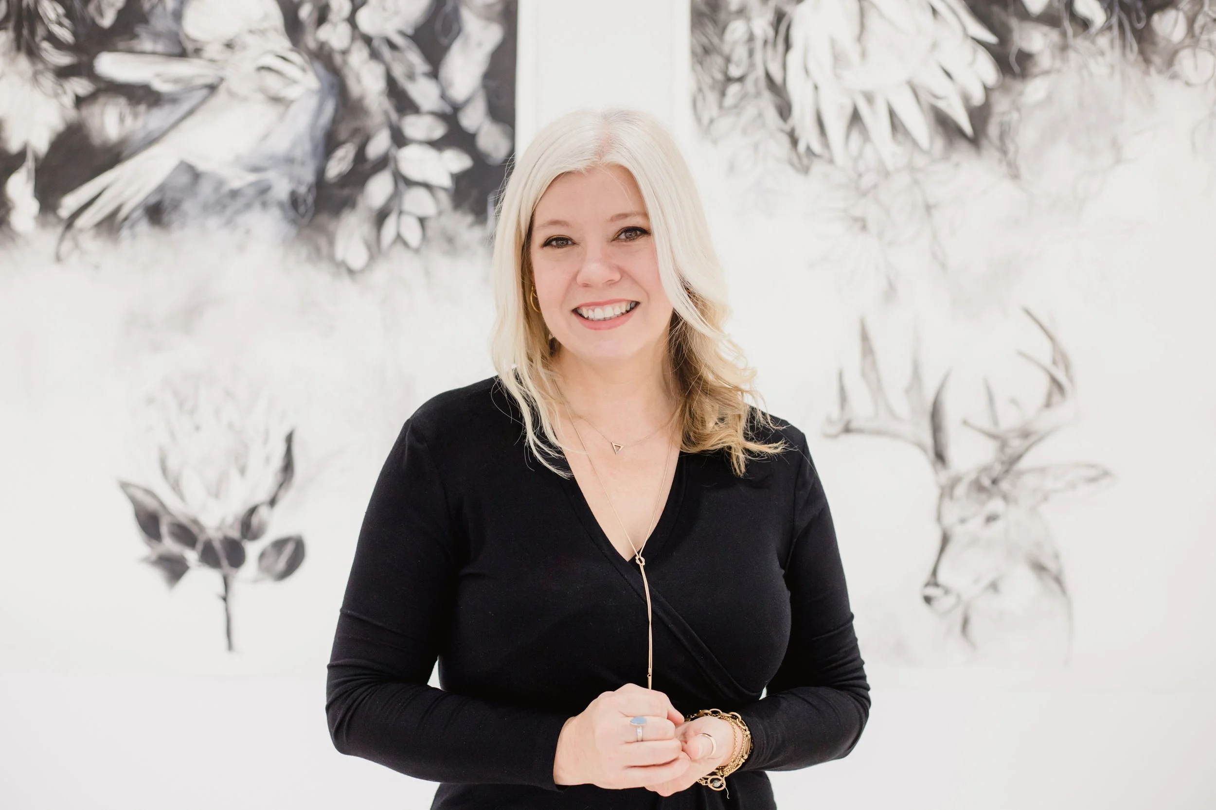 A woman with long blond hair smiling in front of black and white floral artwork.