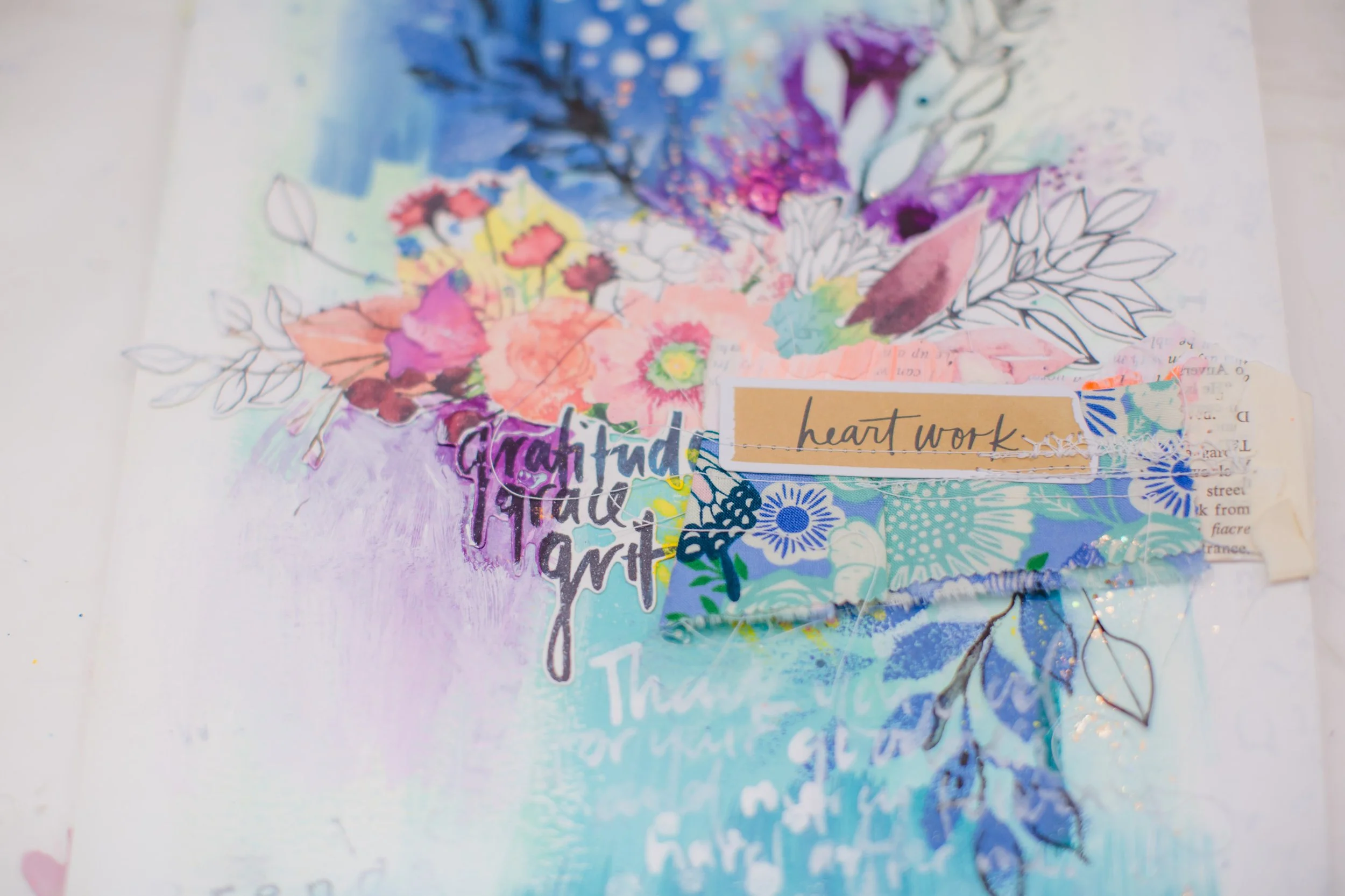 A colorful handmade collage art piece featuring floral illustrations, handwritten words such as 'gratitude' and 'heart work', and layered paper elements.
