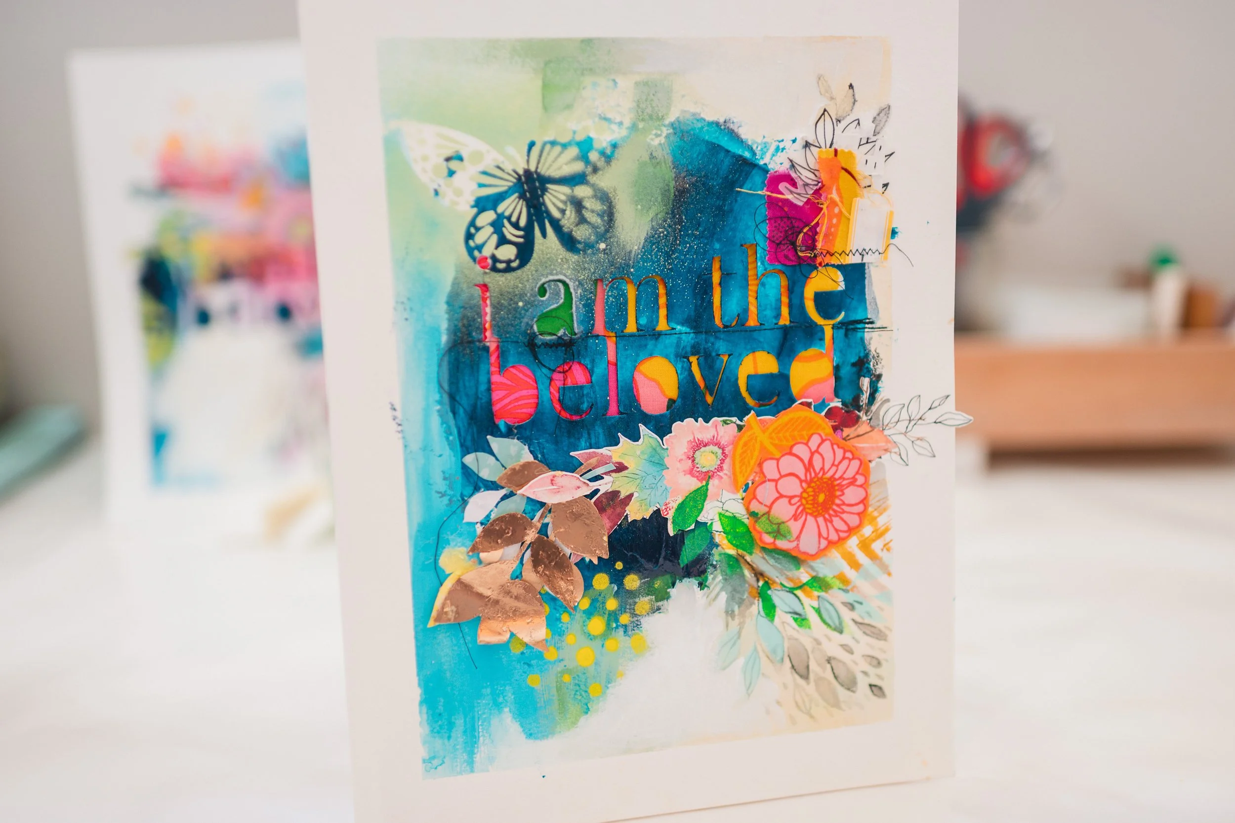 Colorful handmade card with watercolor background, butterfly cutout, and layered paper flowers, with the text 'lam the beloved' in various colors.