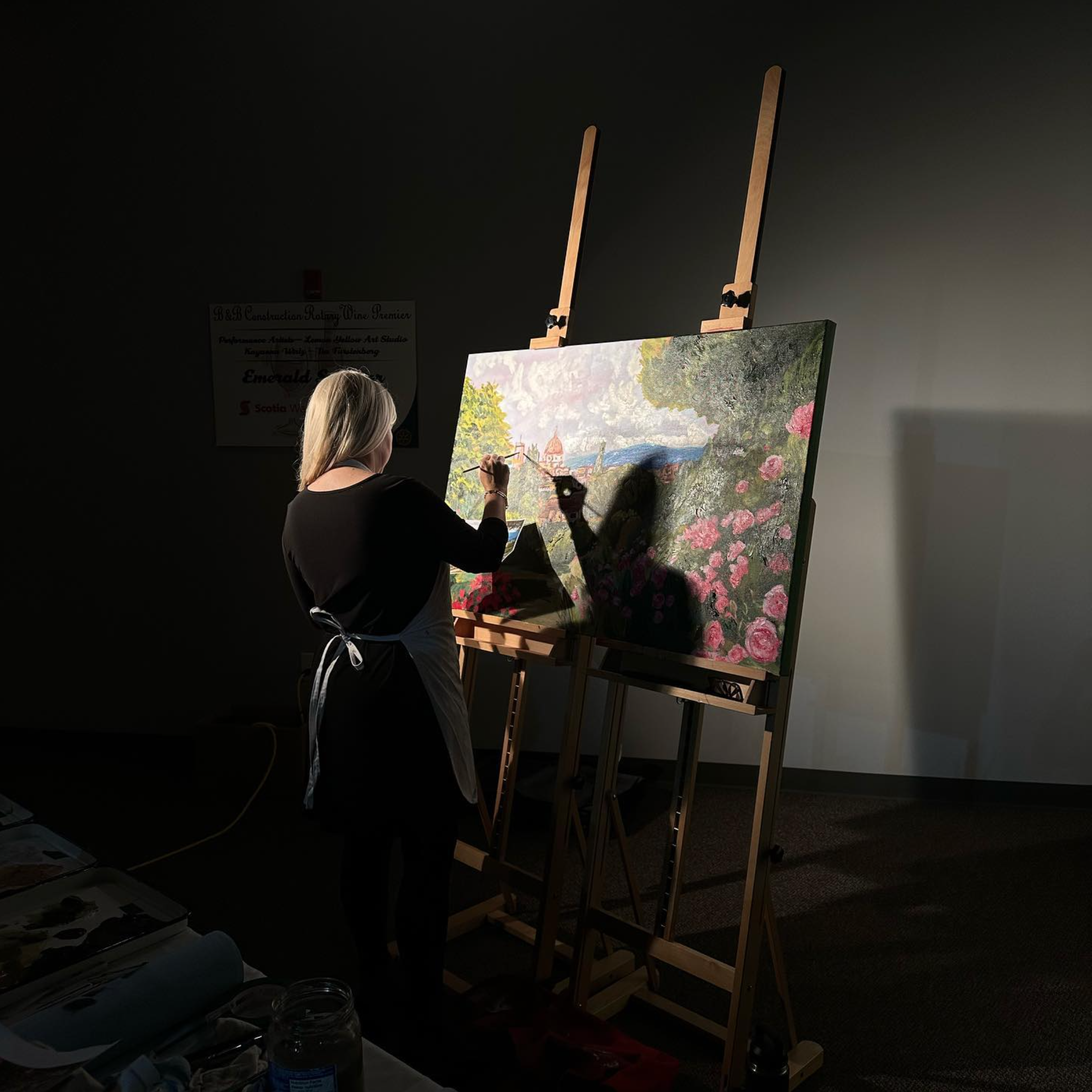 An artist painting a landscape scene on a canvas while standing in a dimly lit room. The artist wears a black top and an apron, with her back to the camera. The canvas is on an easel, and various art supplies, including jars and brushes, are visible on a table nearby.