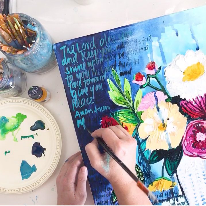 Person painting a floral watercolor in a sketchbook with a Bible verse in the background, surrounded by jars of paintbrushes and paint.