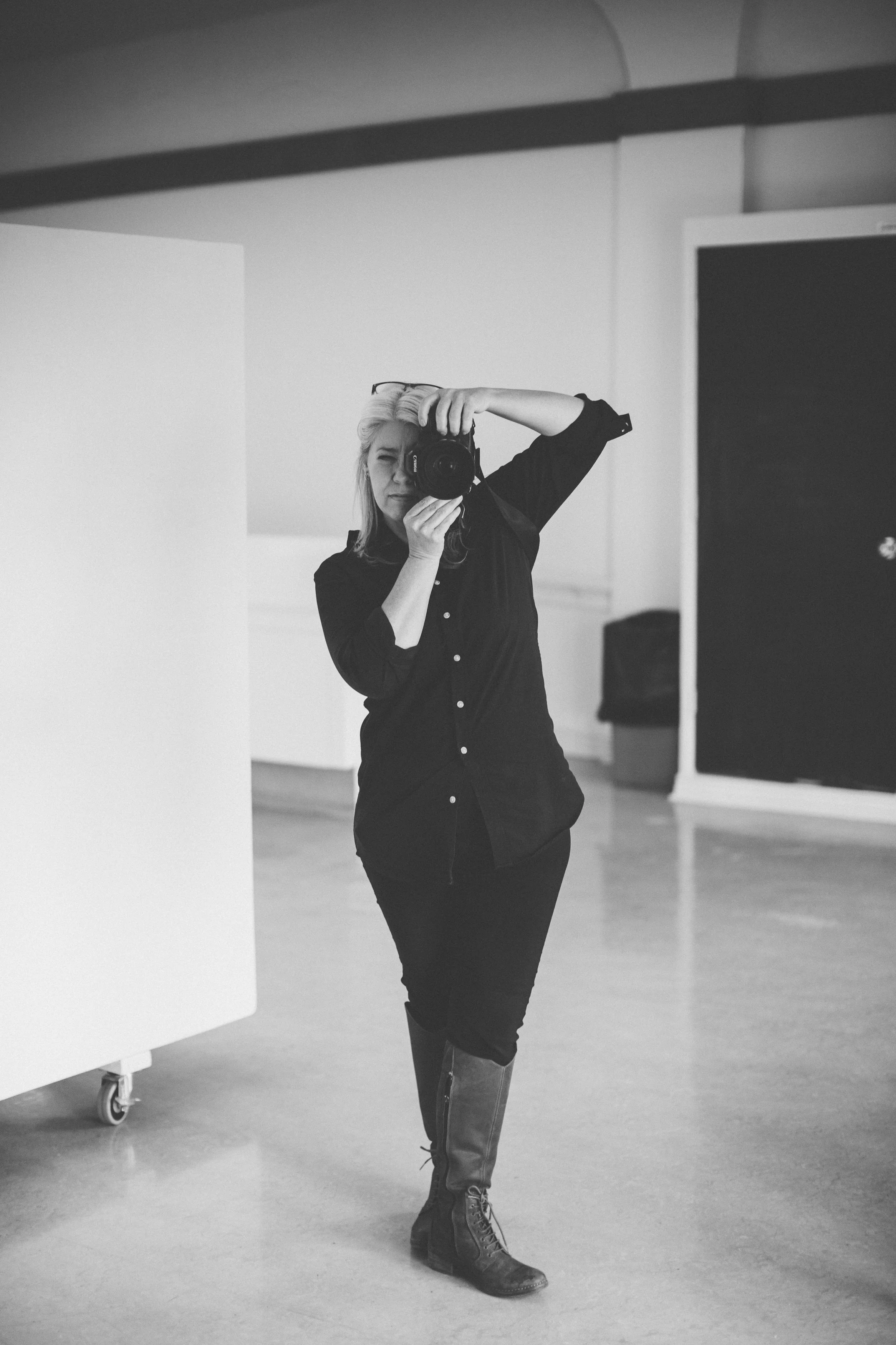 Black and white photo of a woman taking a selfie in a mirror, standing in a room with minimal decor, wearing a black shirt and tall boots, with a camera in hand.