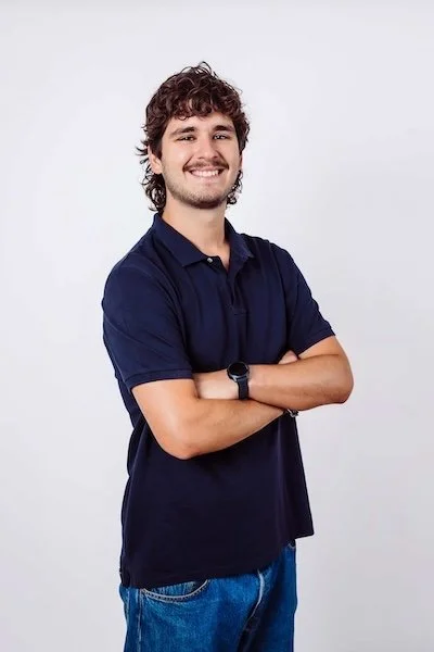 Ein junger Mann mit braunen, lockigen Haaren, blauer Polohemd und blauen Jeans, der vor einem weißen Hintergrund lächelnd steht, mit verschränkten Armen und einem Handgelenk-Uhr.