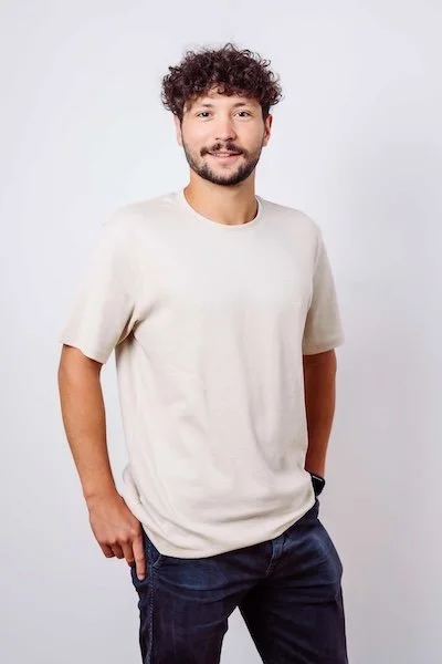 Ein junger Mann mit lockigem Haar und Bart, trägt ein beiges T-Shirt und dunkle Jeans, steht vor einem weißen Hintergrund.