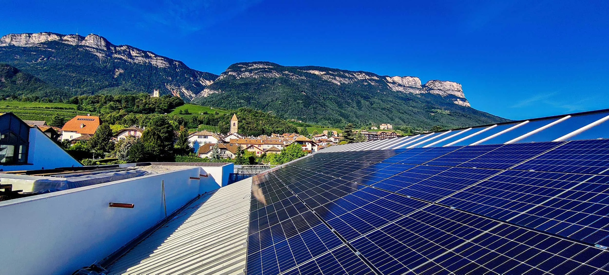 Dach mit Solarpaneelen vor einer malerischen Stadt mit Kirchturm und grünen Hügeln, im Hintergrund Berge bei klarem Himmel.
