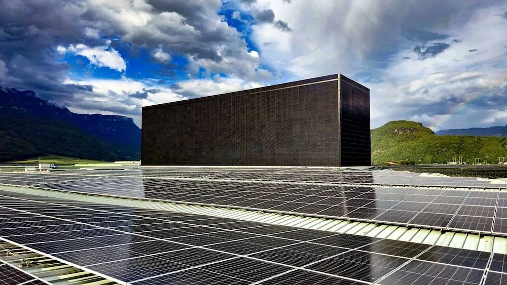 Solarpanels vor einem schwarzen Gebäudeblock, mit Bergen und Himmel im Hintergrund.