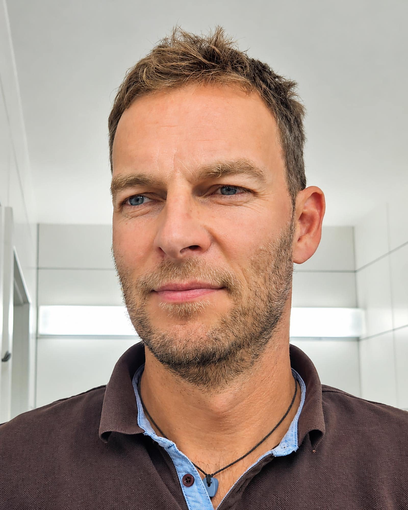 Nahaufnahme eines Mannes mit blauen Augen, braunem Haar und Bart, im Hintergrund eine weiße Wand.