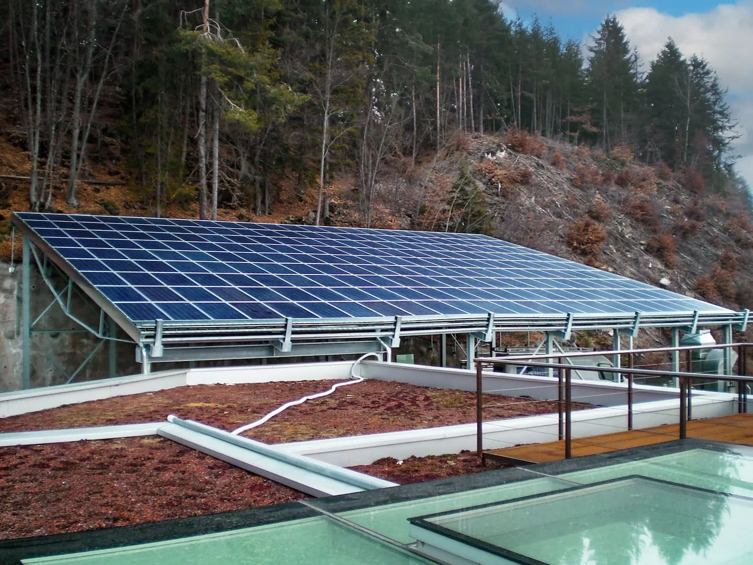 Dachanlage mit Solarmodulen auf einem Flachdach vor einem Wald mit Bäumen auf einem Hügel.