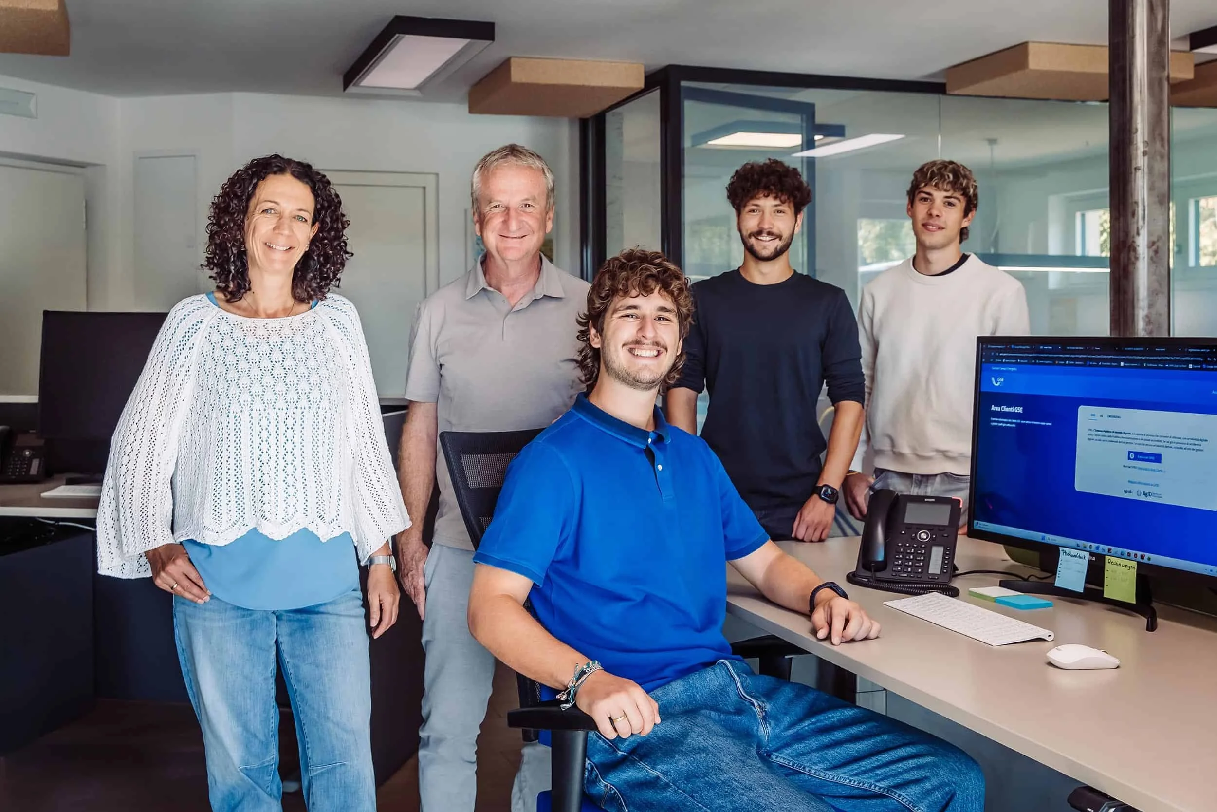 Team Wenter Service im modernen Büro am Ritten, drei Männer und zwei Frauen, die vor einem Computer sitzen und stehen, lächelnd in die Kamera schauen.