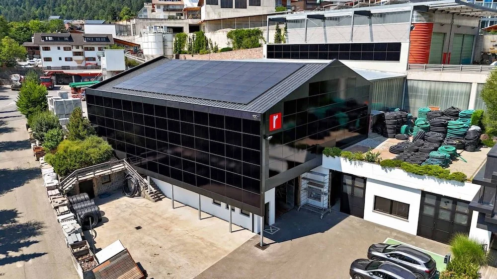 Luftaufnahme eines modernen Gebäudes mit schwarzer Glasfassade und Solarzellen auf dem Dach, umgeben von Reifenlagern, Autos und begrünten Flächen.