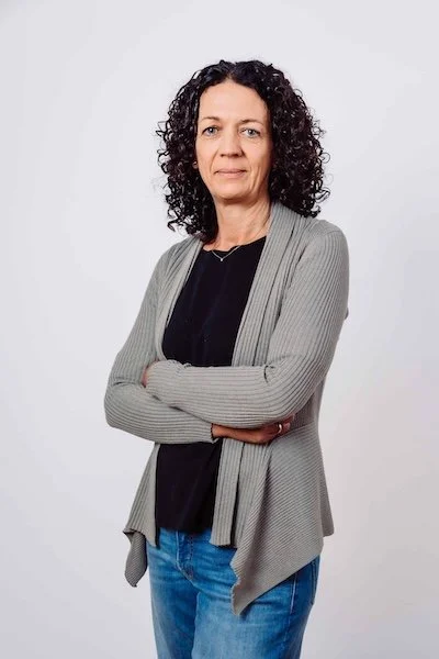 Eine Frau mit schwarzen, lockigen Haaren, die einen grauen Cardigan, ein schwarzes Top und Jeans trägt, steht vor weißem Hintergrund, die Arme verschränkt.
