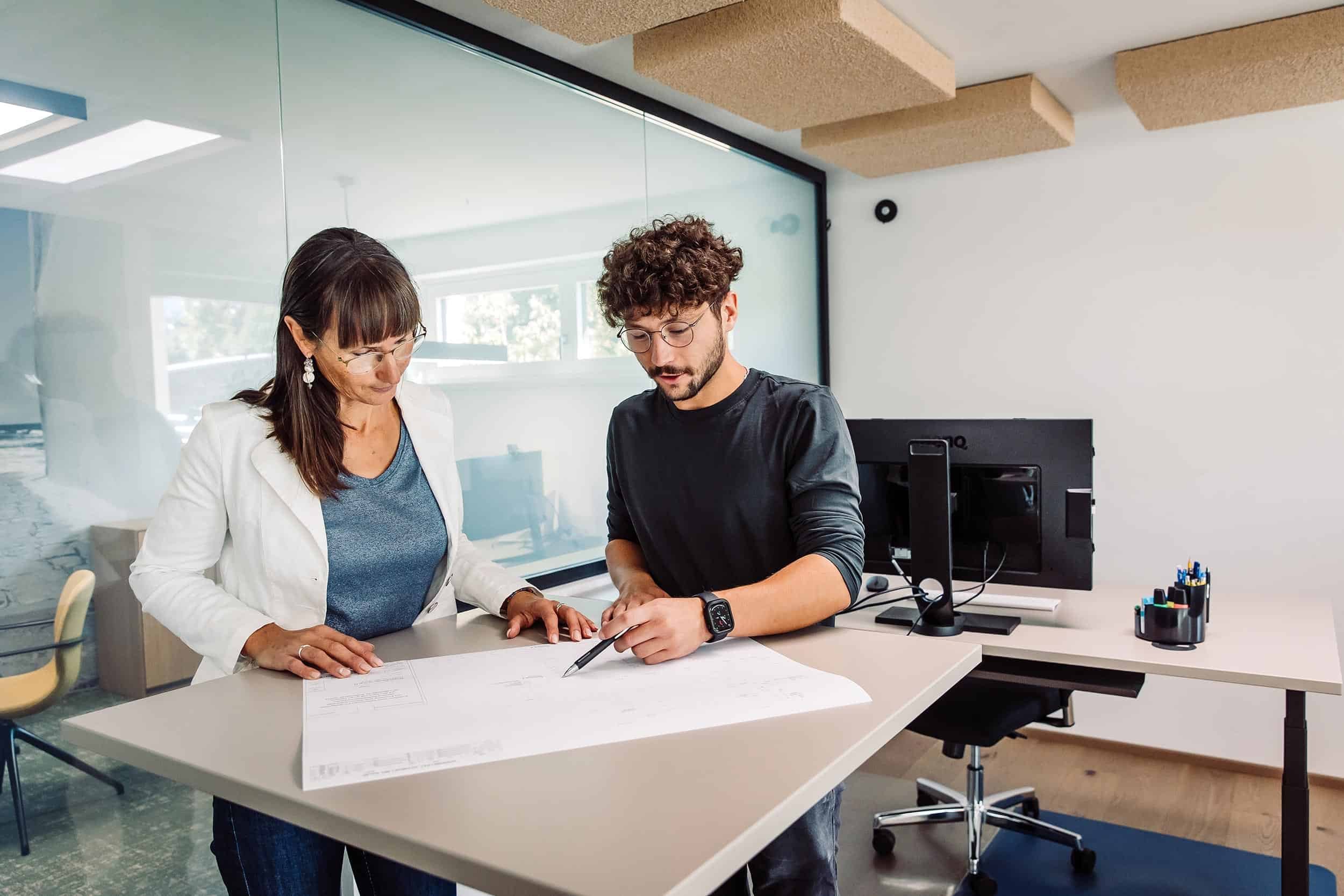 Zwei Menschen, eine Frau und ein Mann, arbeiten zusammen an einem Dokument auf einem Tisch in einem modernen Büro, während sie sich austauschen.