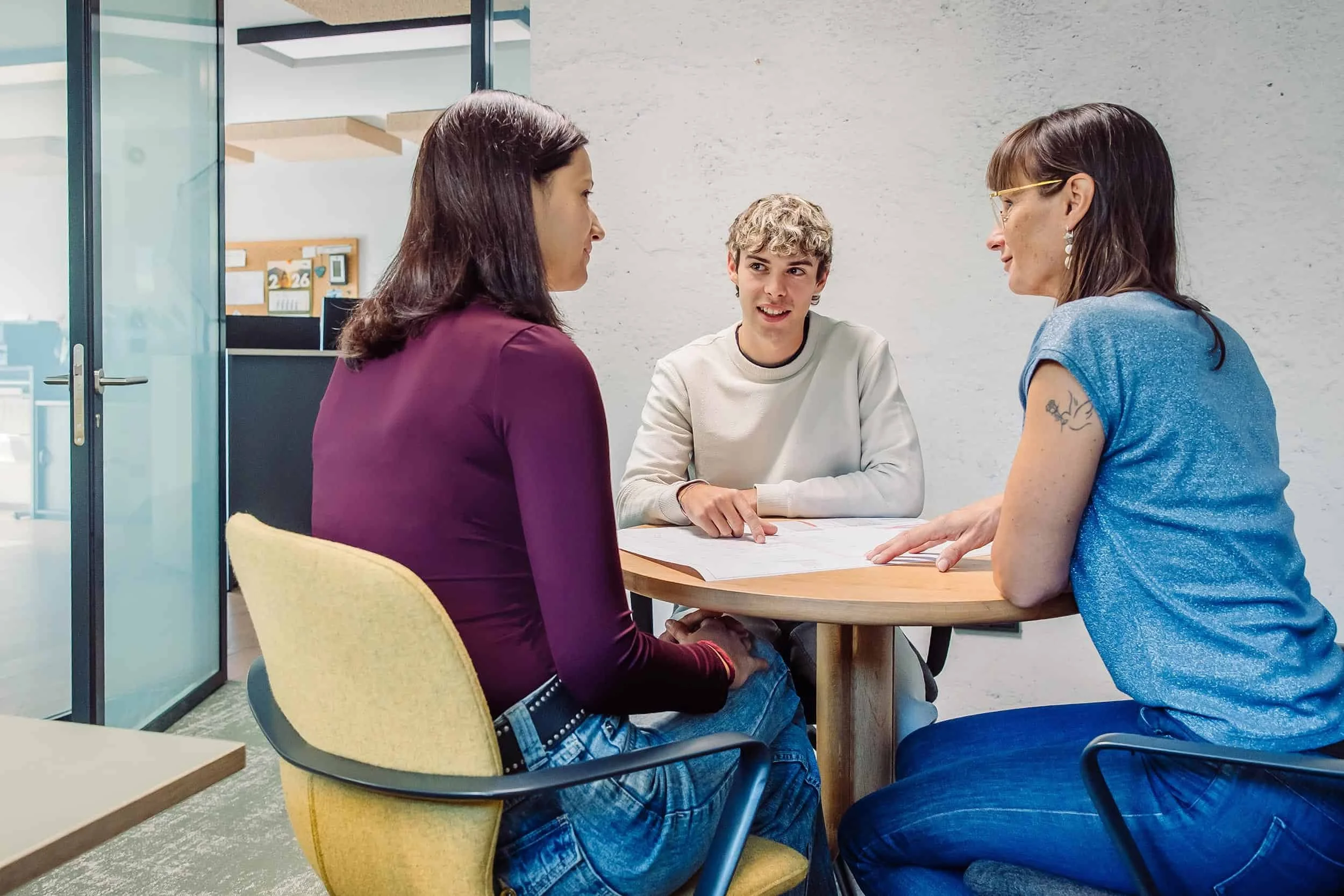 Drei Menschen sitzen um einen runden Tisch und führen ein Gespräch. Zwei Frauen und ein junger Mann.