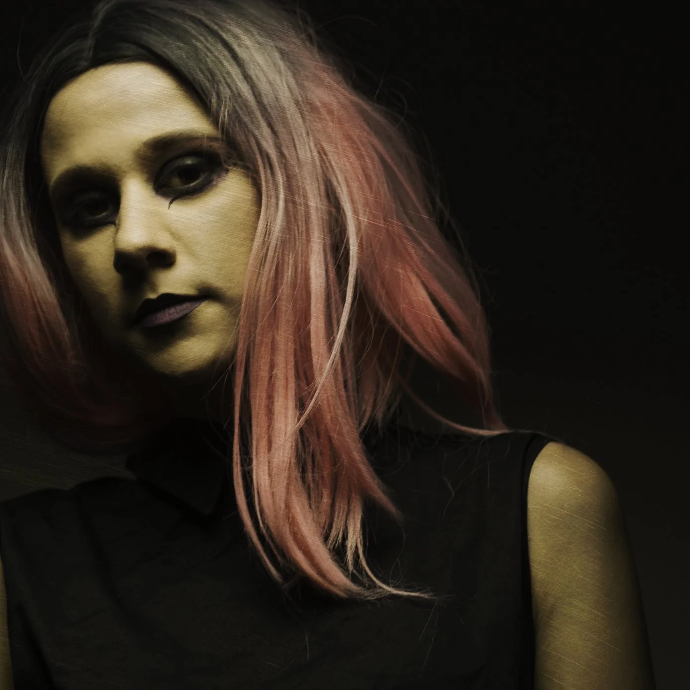 A woman with medium skin tone and pink hair with black roots, wearing dark makeup, a black sleeveless top, looking slightly to the side against a dark background.