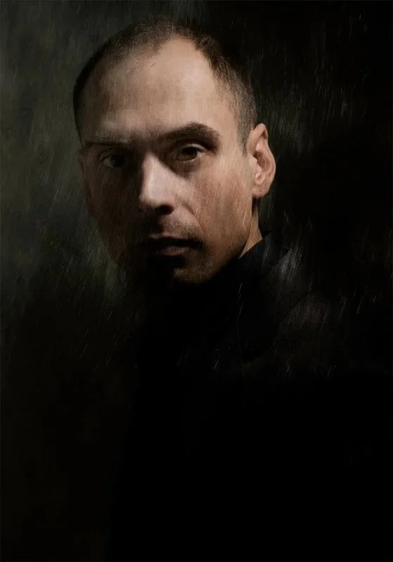 A close portrait of a young man in a vey dark space. He is lit on one side with a single light. Texture is laid over the image.
