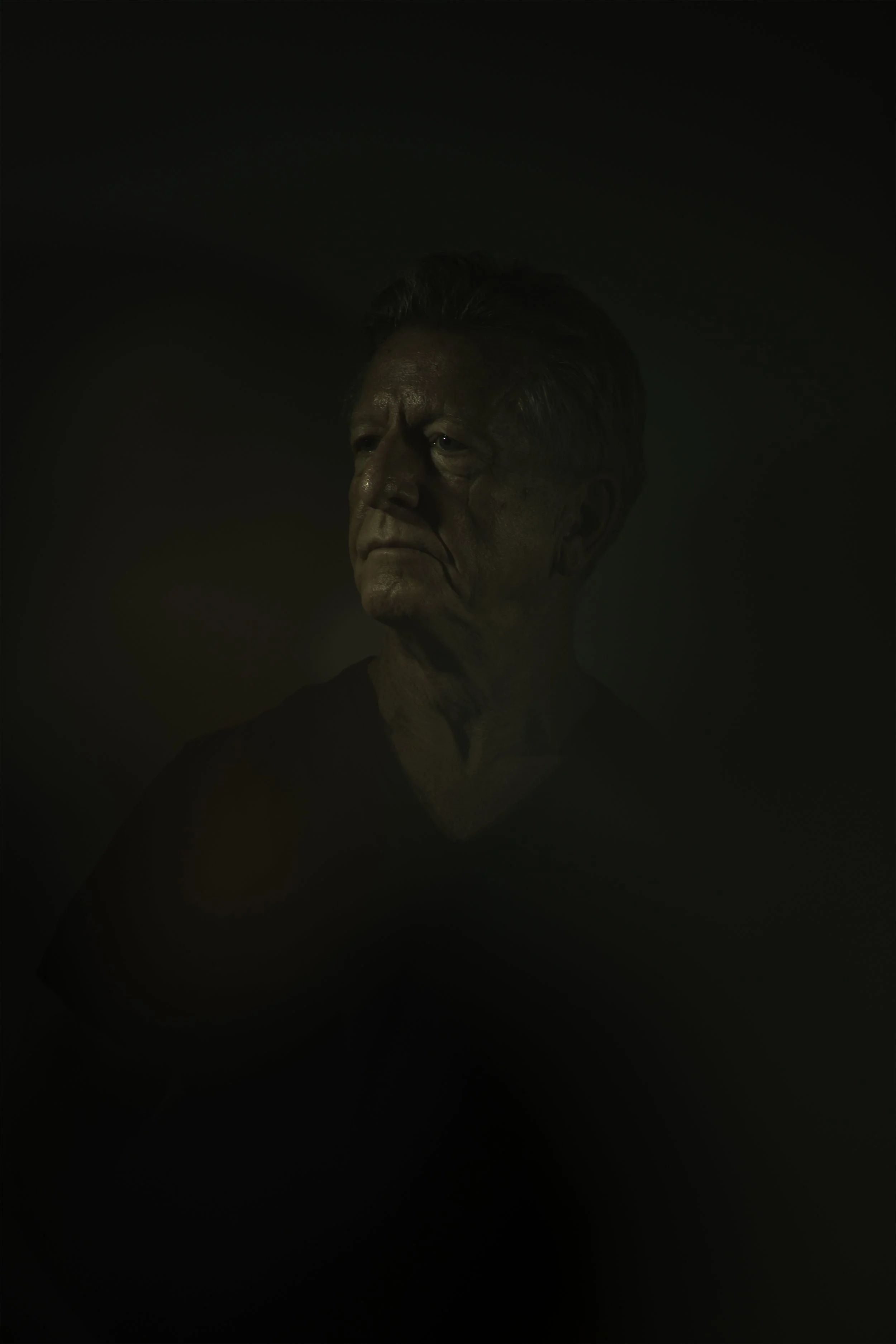 A close-up portrait of an elderly man with short, gray hair and a serious expression, set against a dark background.