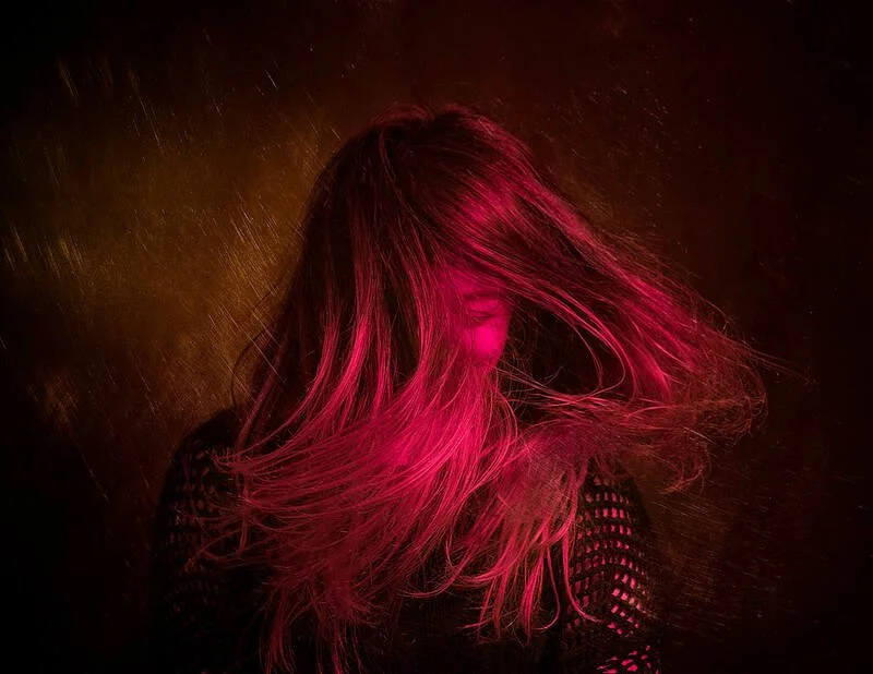 A dark portrait of a person with plenty of hair covering their face. They are lit with a red light.
