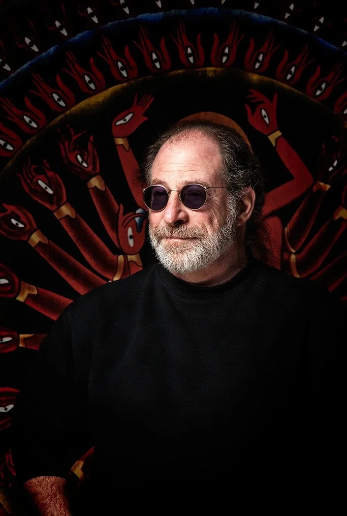 Portrait of a bespectacled man in front of a tapestry.
