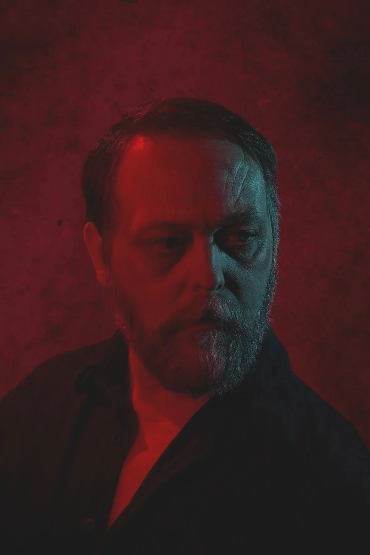 A man with a beard and mustache looking directly at the camera against a red background. The lighting is dark with red highlights.