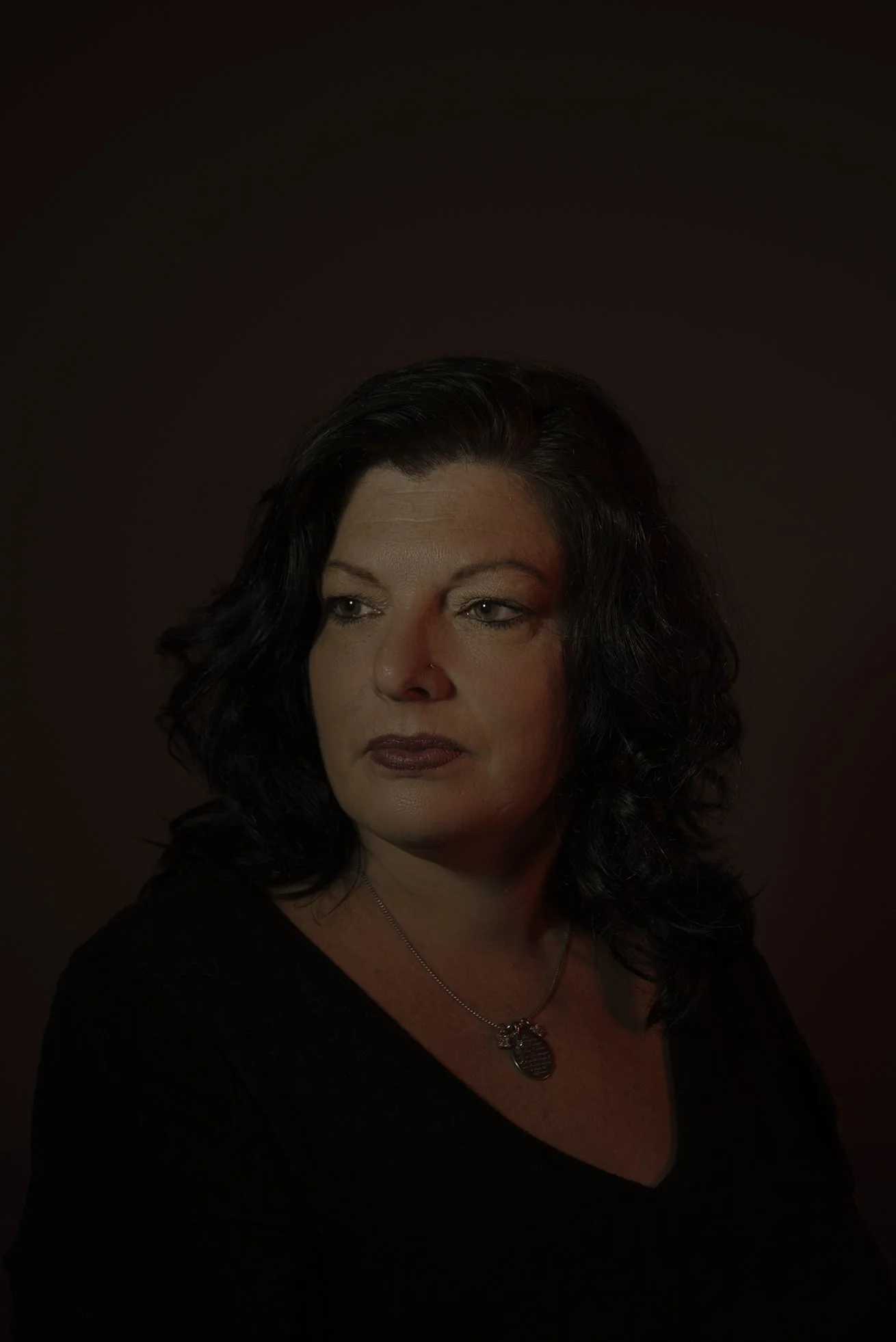 A woman with dark, curly hair, wearing a black top and a silver necklace, looking slightly to the side against a dark background with red lighting on her face.
