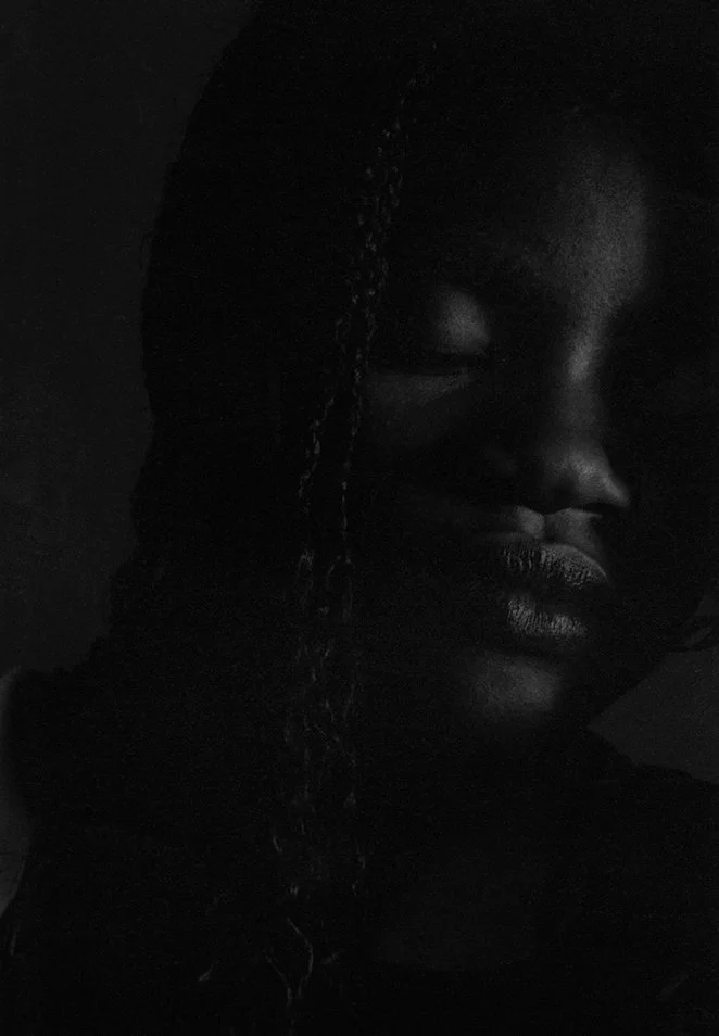 A monochromatic portrait of a dark skinned woman in a black room.