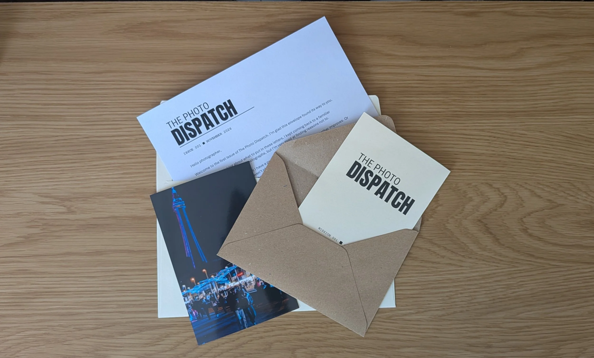 Photo of various printed materials including a postcard/letter titled 'The Photo Dispatch,' greeting card, and a glossy photograph of a cityscape at night with blue neon lights on a wooden surface.