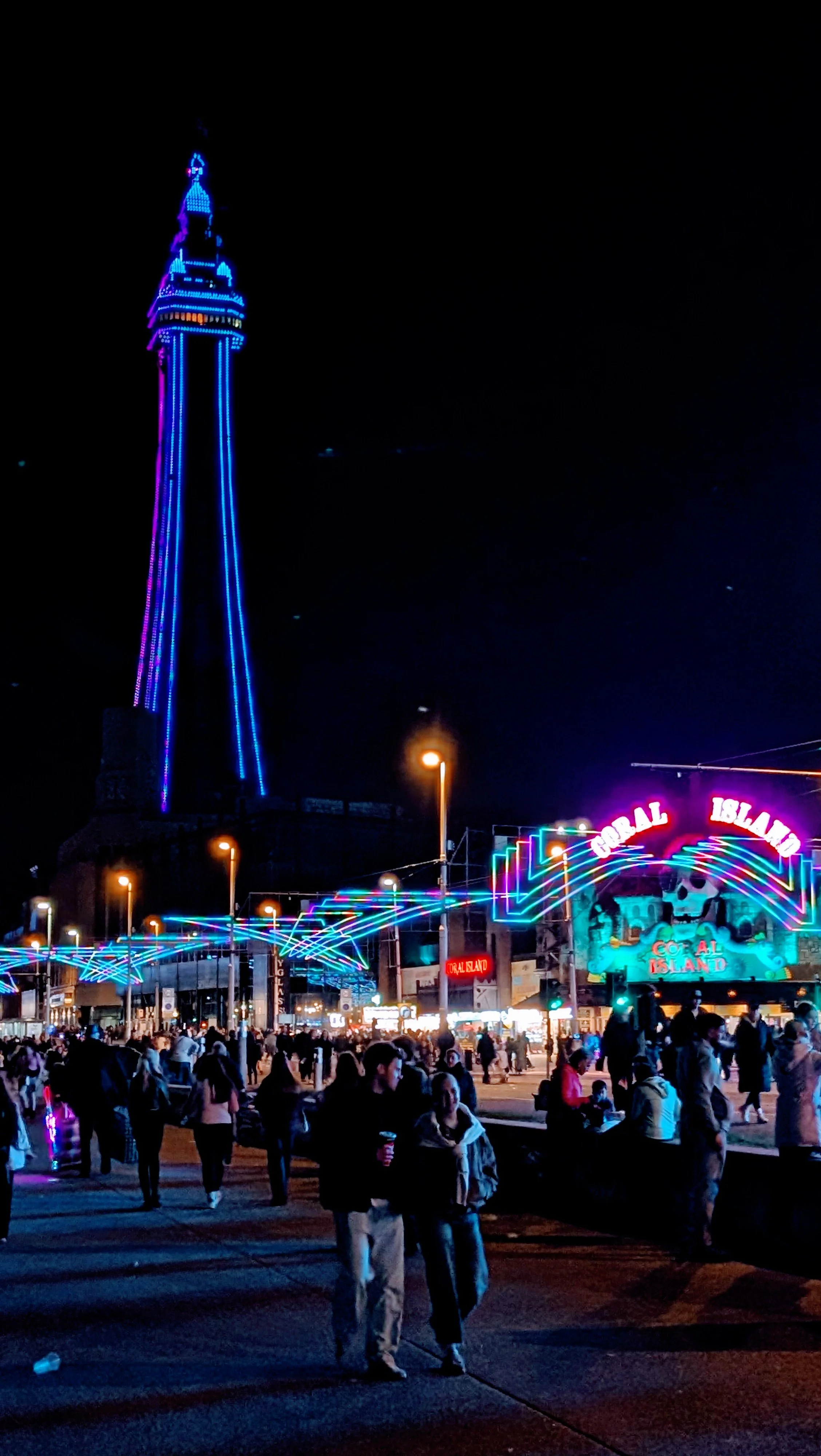Crowd of people at a night fair or carnival, with a brightly lit tower and colorful neon lights in the background, including the sign 'Coral Island'.