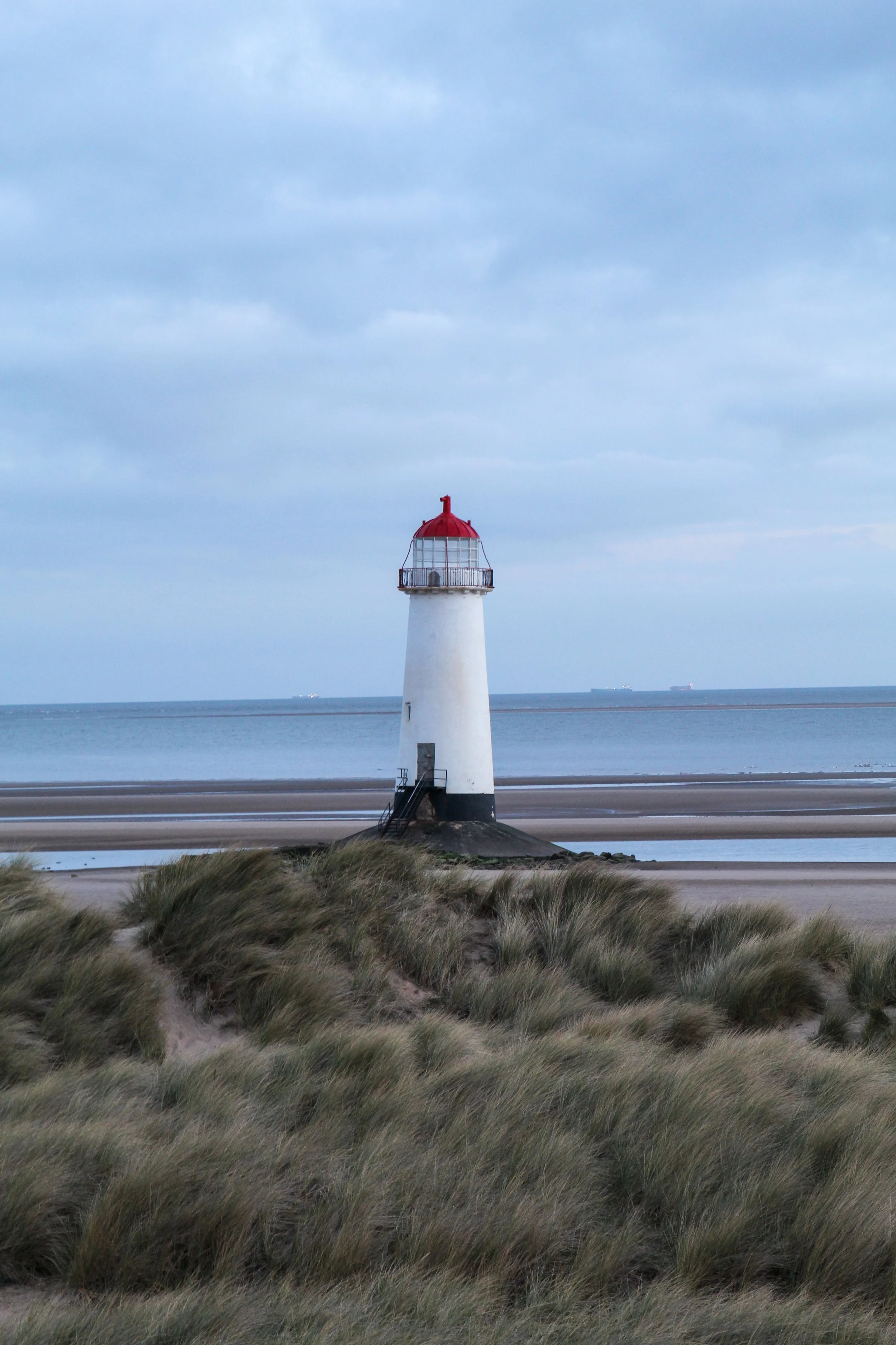 A white lighthouse with a red top standing near the ocean, with grassy dunes in the foreground and ships on the water in the background under a cloudy sky.