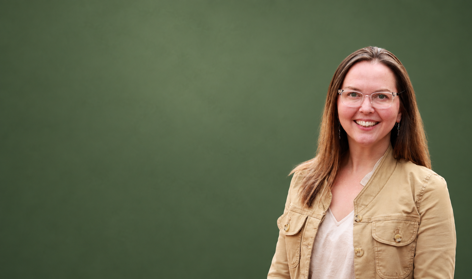 A woman with long brown hair, glasses, and earrings, smiling, wearing a tan jacket over a white shirt, standing in front of a green background.