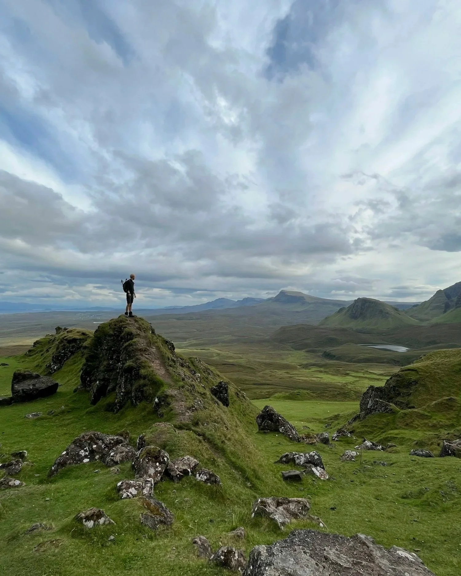 A lone hiker standing on a rocky hillside overlooking a vast green valley with mountains in the distance under a cloudy sky.
