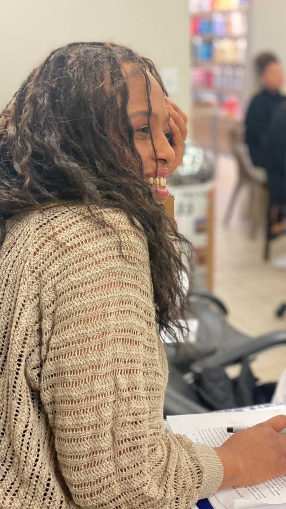 A woman with curly hair smiling while sitting at a table, holding a pen and reading a book or magazine. In the background, a blurred person is standing in a bookstore or library.