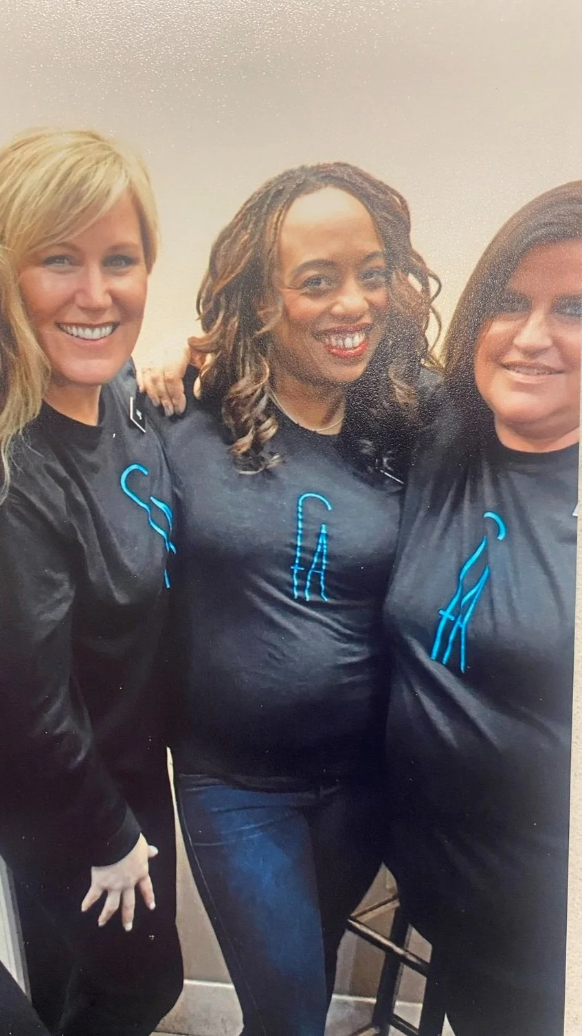 Three women smiling and wearing black t-shirts with blue LED-style designs, standing close together in an indoor setting.