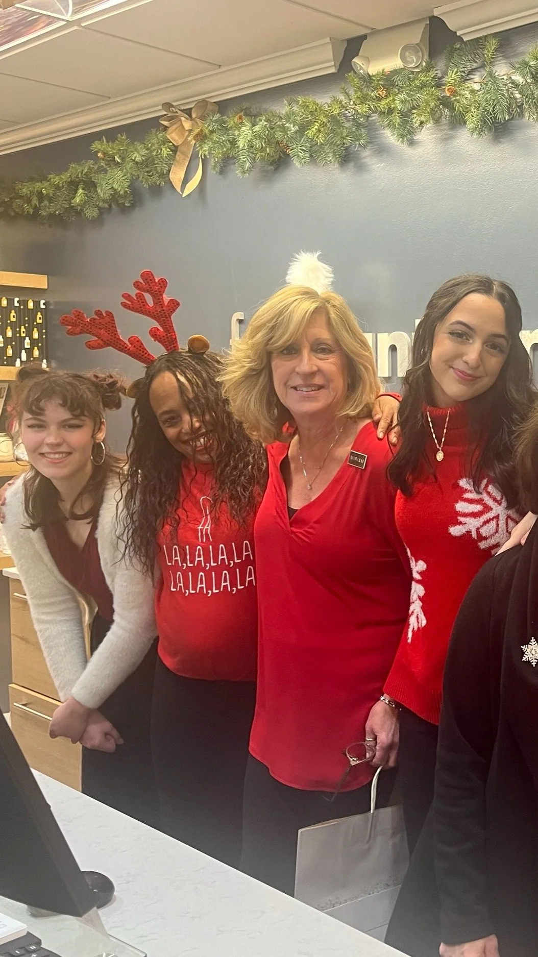 Four women celebrating Christmas, wearing festive sweaters and accessories, standing close together indoors near a decorated wall with greenery and a bow.