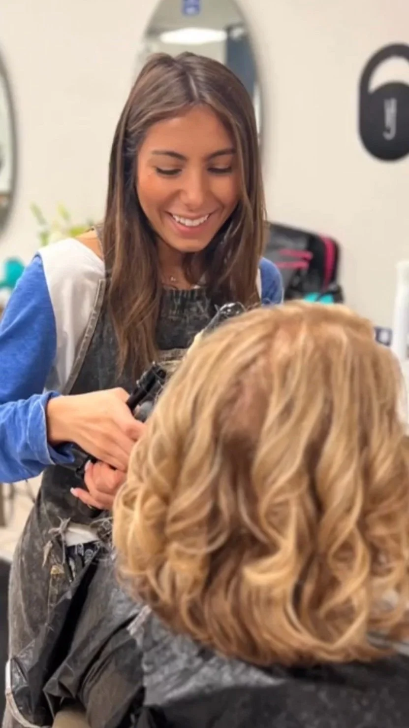 A hairstylist smiling while working on a customer in a salon, with hair styling tools and products visible in the background.