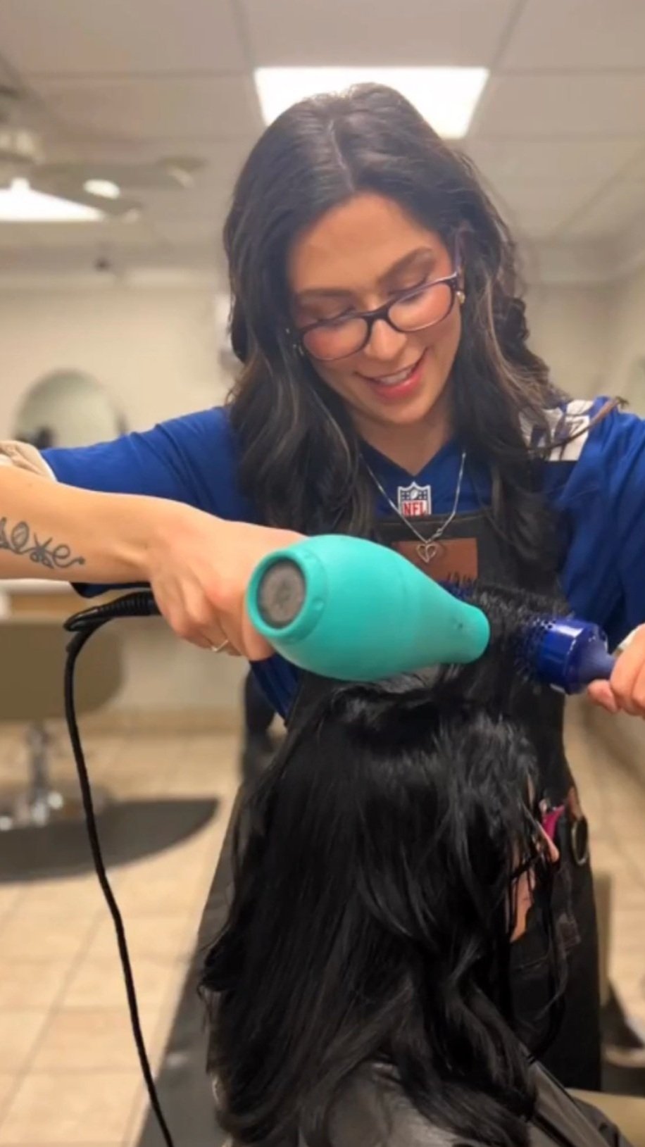 A female hairdresser with glasses blow-drying a client's long black hair in a salon.