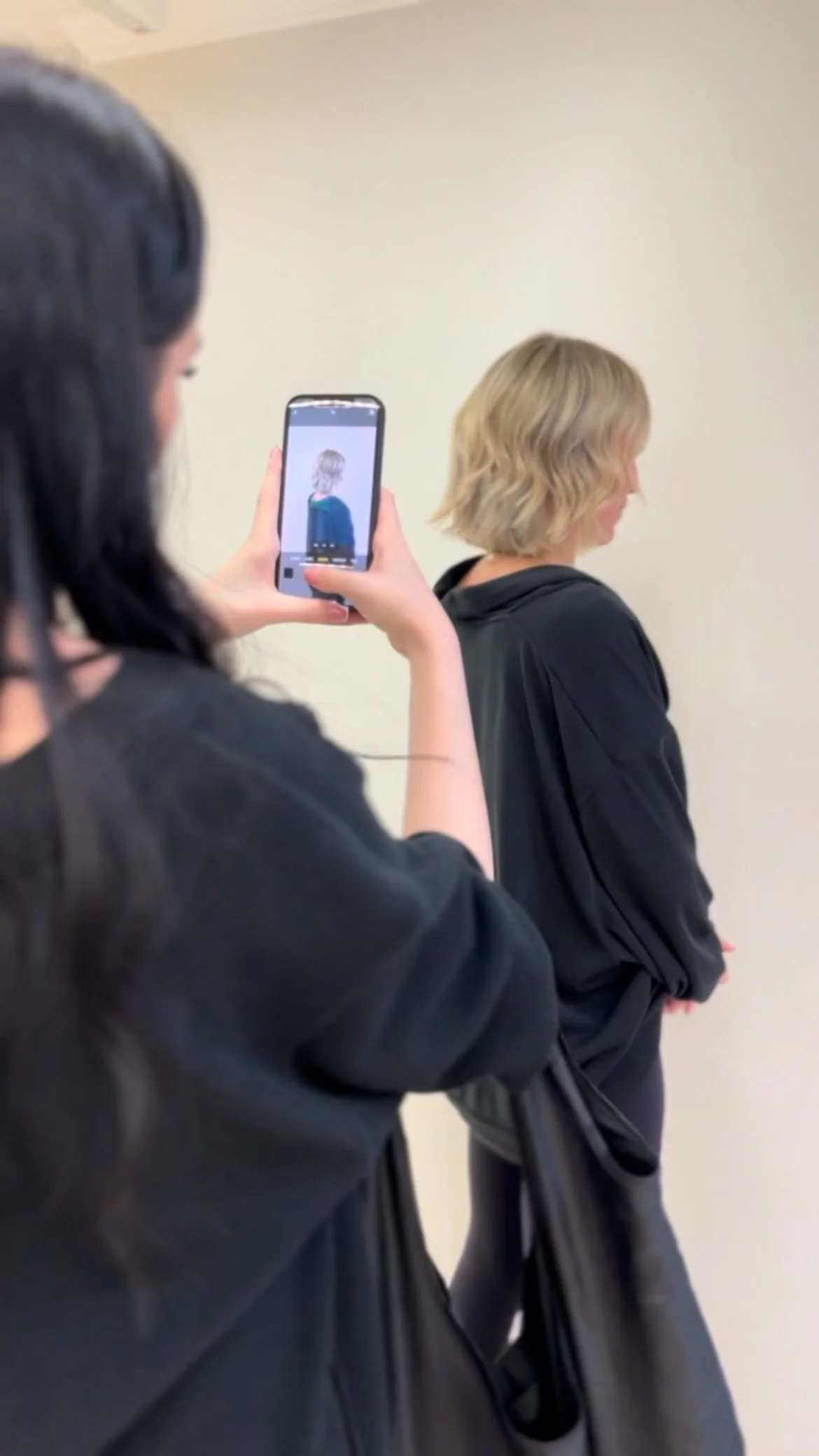 A person with dark hair taking a photo of a woman with blonde hair in a black hoodie against a plain beige wall.