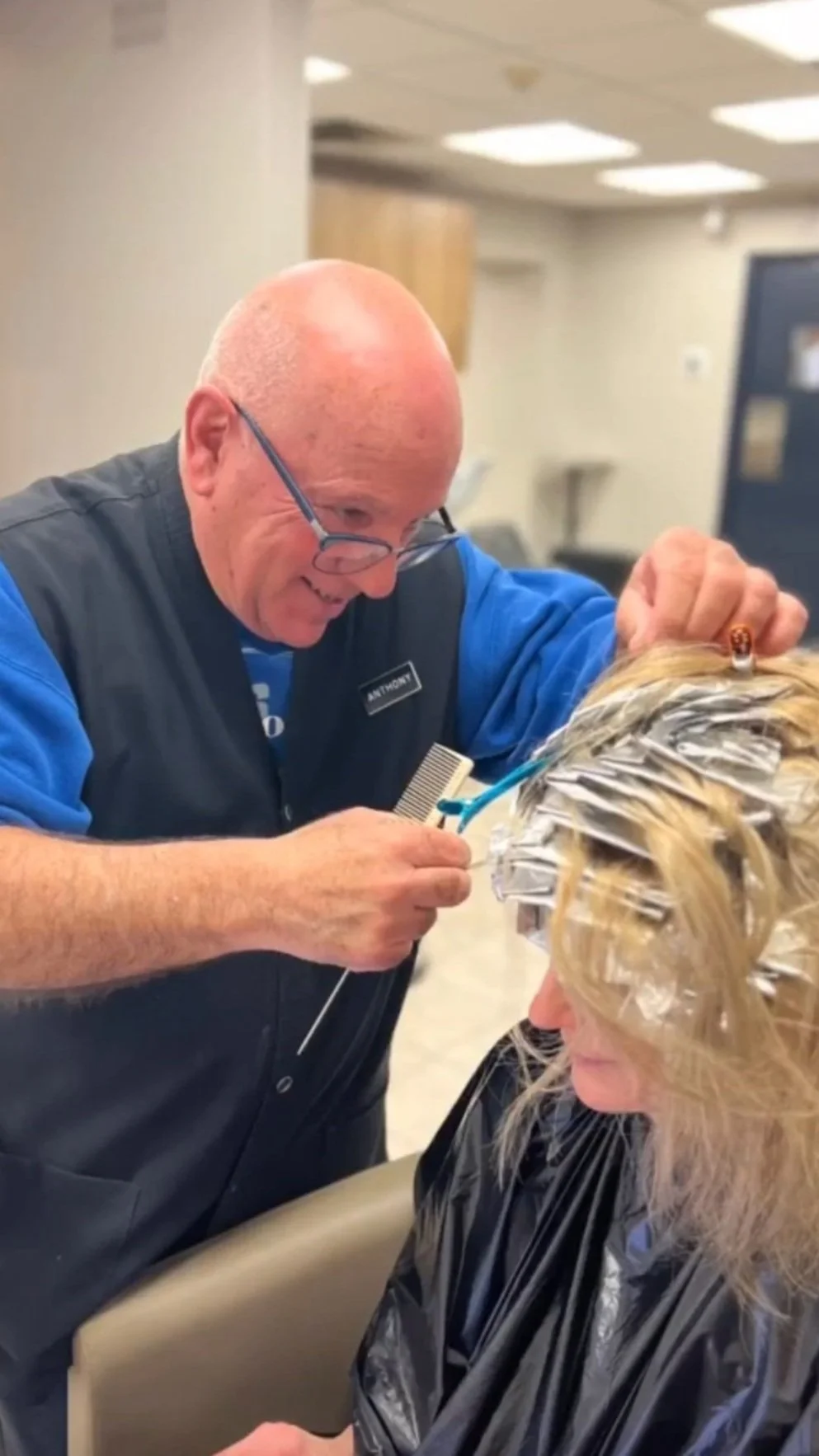 A man with a bald head and glasses is giving a haircut to a woman whose hair is covered with foil hair rollers. The man is smiling and using a hairbrush, while the woman is seated in a salon chair.
