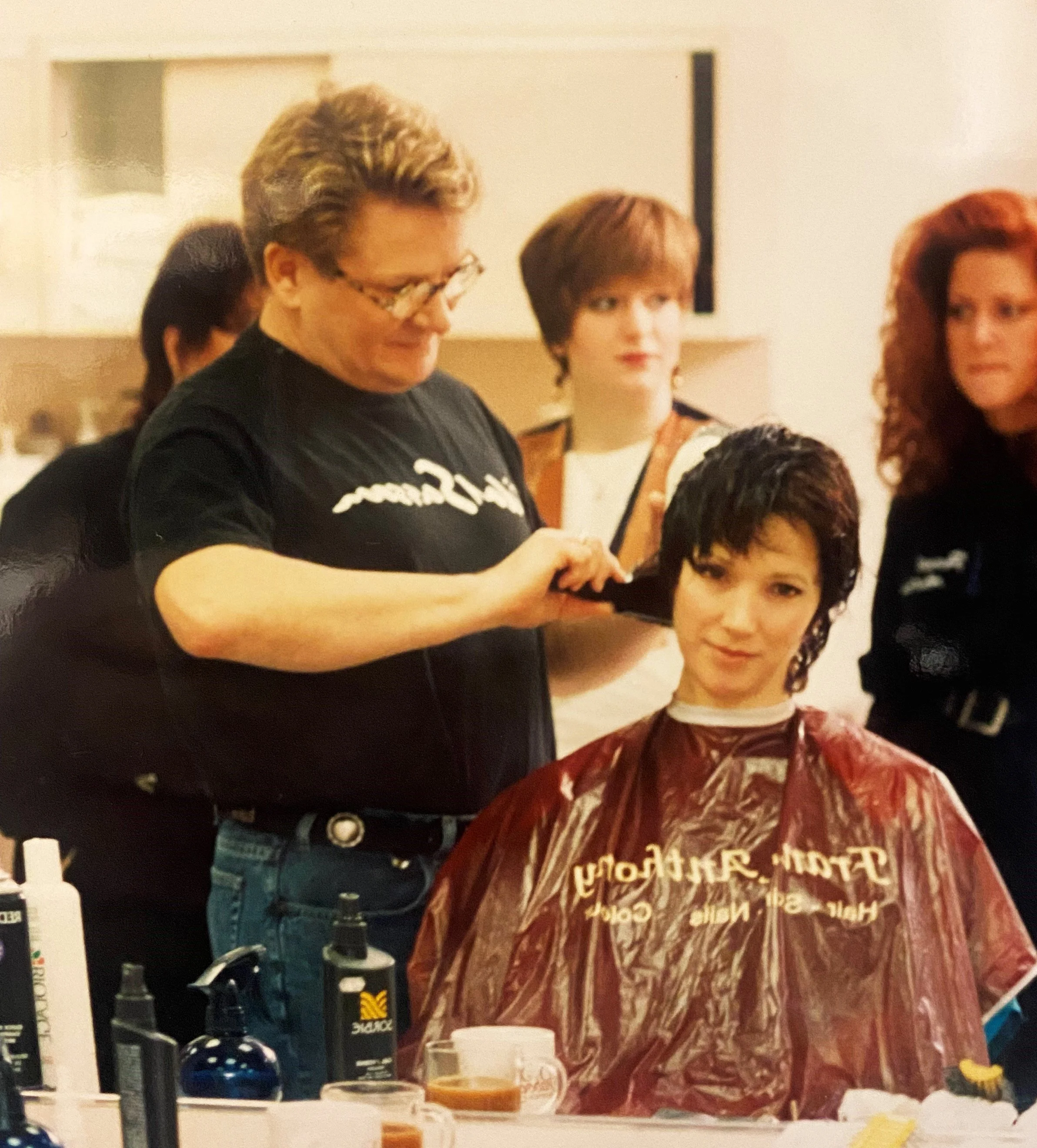 A woman with short, curly hair wearing a red and brown patterned shirt sits at a table, with a person who has short, curly, dark hair and is wearing a dark shirt standing behind her. The table has various bottles, cups, and containers. In the background, there are two women with short hair, and the room appears to be indoors with beige walls and cupboards.