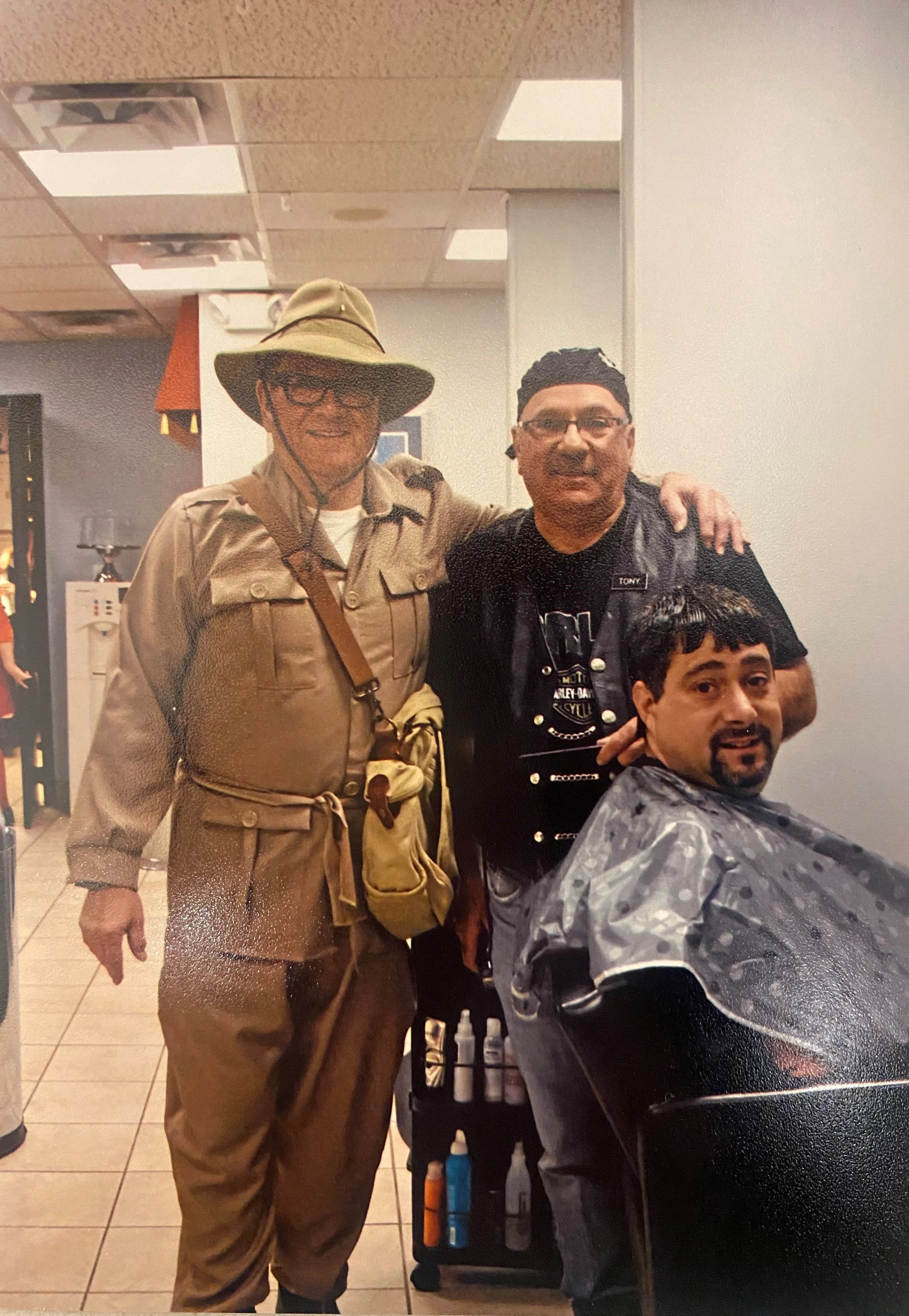 A man in a khaki outfit and a broad-brimmed hat standing in a barber shop with arm around a barber wearing glasses and a bandana, with chairs and barber supplies in the background.