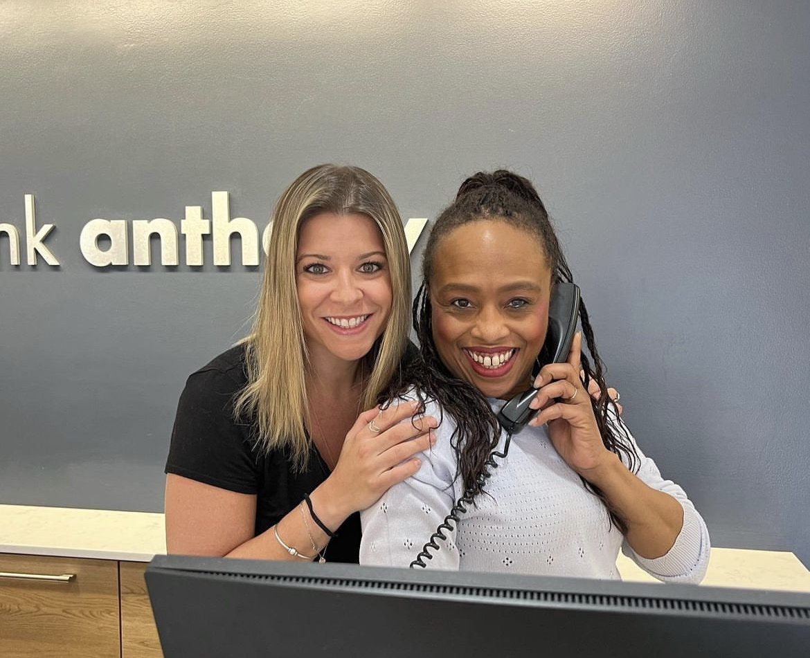 Two women smiling at the camera at a reception desk. One woman is on the phone and the other is standing close, with her hand on the first woman's shoulder. Behind them is a gray wall with the text 'frank anthony' partially visible.