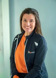 Smiling woman with long brown hair wearing a navy sports jacket and an orange shirt inside, standing indoors with a light green background.
