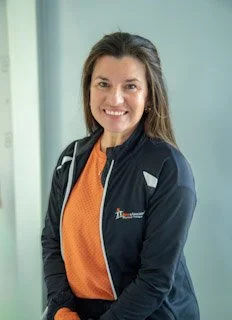A smiling woman with long brown hair wearing an orange shirt and a black jacket standing indoors.