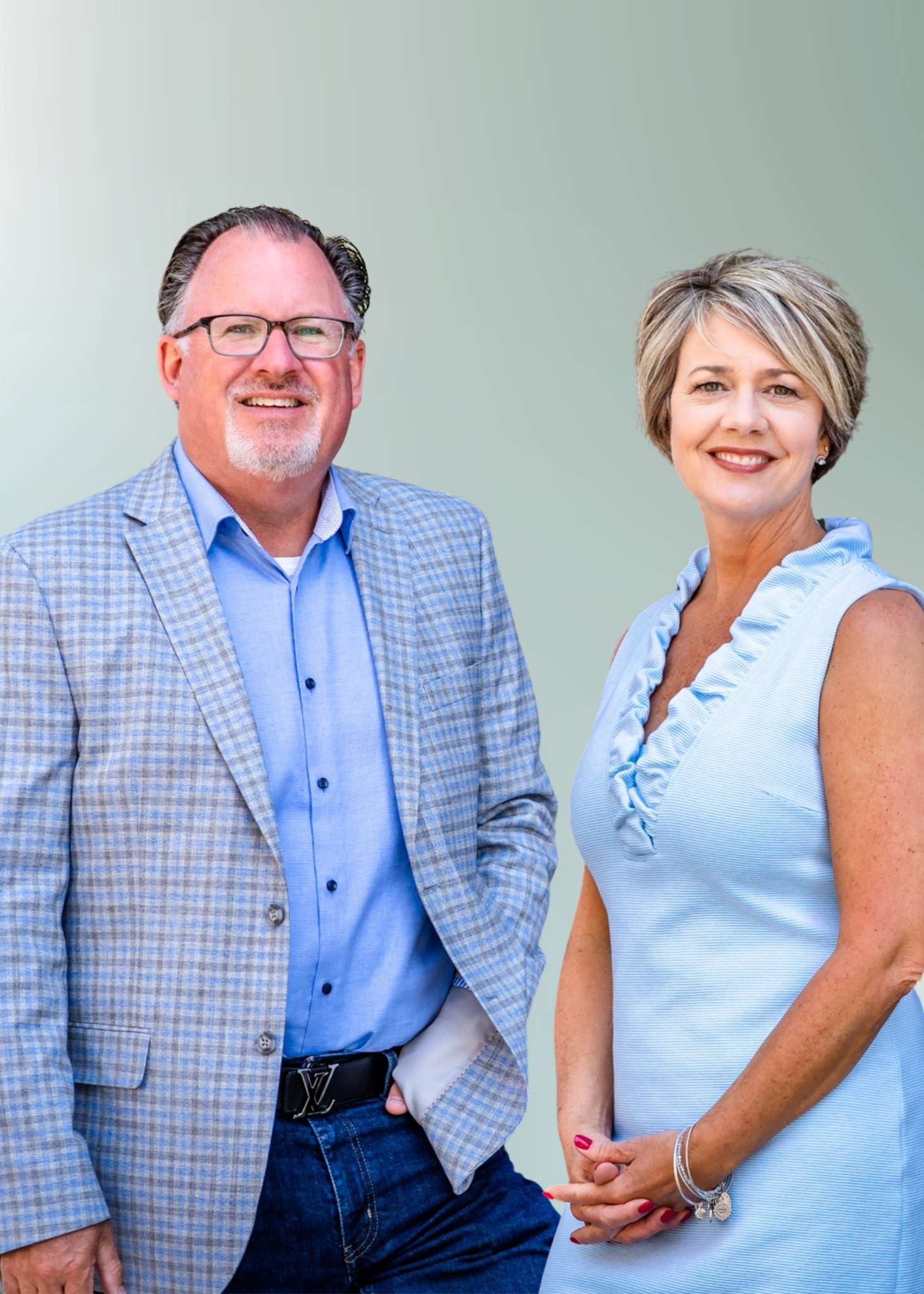 A man wearing glasses and a plaid blazer is smiling and standing next to a woman in a sleeveless blue dress. They are posing against a plain, light green background.