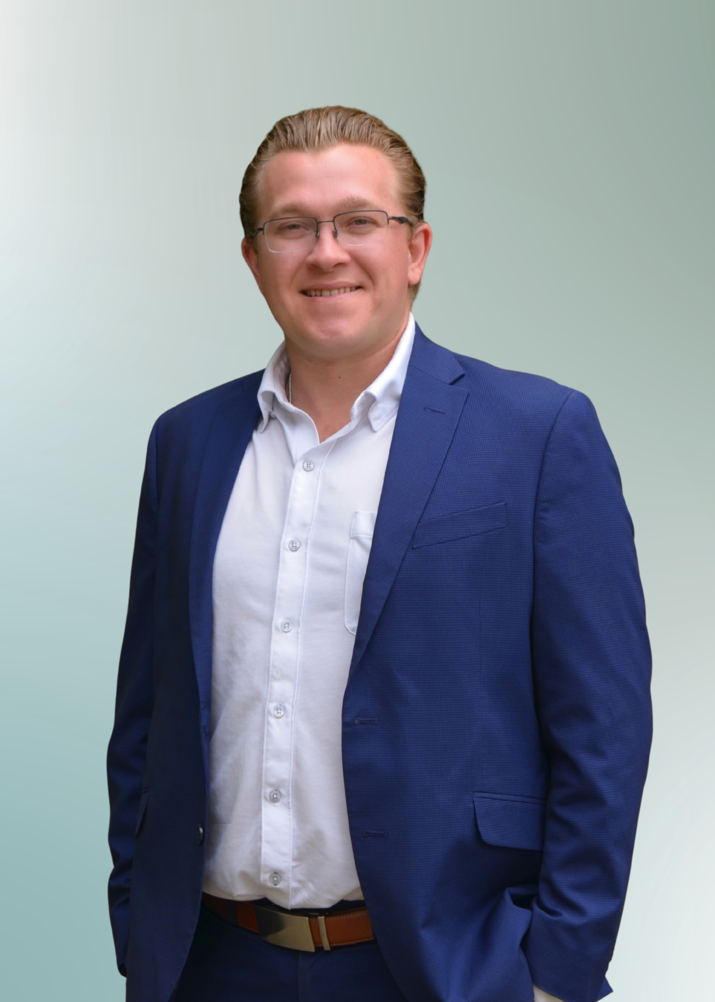 A young man with light skin, wearing glasses, a white dress shirt, a blue suit jacket, and brown belt, smiling at the camera against a gradient green background.